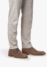 Botas de ante marrón hasta el tobillo con cordones, que presentan un tacón de cuero marrón claro en contraste y detalles texturizados, combinadas con pantalones cuadros gris claro.
