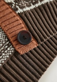 Detailní záběr hnědého a béžového pleteného materiálu s žebrovanou texturou a velkým tmavým knoflíkem na rezavě zbarveném pleteném pásku.