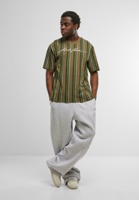 Olīvu zaļš svītrots t-krekls ar īsām piedurknēm un baltu paraksta detaļu, kombinēts ar brīviem pelēkiem sporta biksēm un bežiem sporta apaviem.