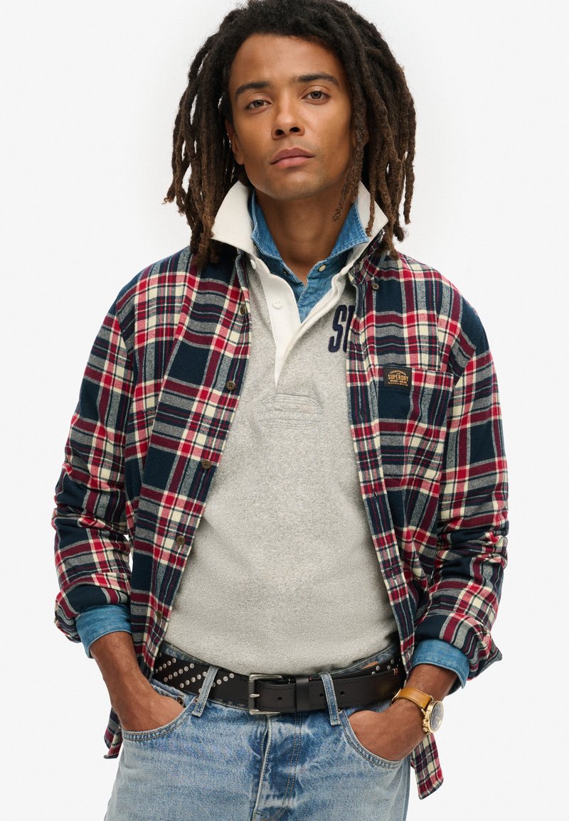 Czerwona, granatowa i biała koszula w kratę z flaneli na szarym polu; dżinsy; czarny pasek z ćwiekami; zegarek na nadgarstku; swobodny, warstwowy wygląd.