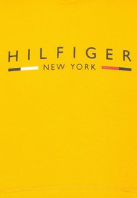 Sudadera amarilla con "HILFIGER NEW YORK" impreso en azul marino. Líneas de acento blancas y rojas debajo del texto. Material suave y texturizado.
