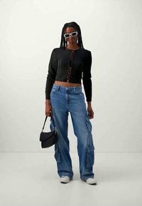 Μαύρο κοντό ζακέτα με κουμπιά μπροστά, ανοιχτό μπλε cargo τζιν με πλαϊνές τσέπες, λευκά γυαλιά ηλίου και μια μικρή μαύρη τσάντα χειρός.