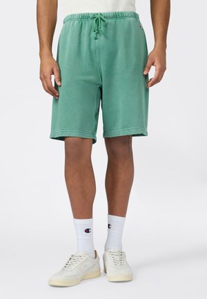 Hombre con pantalones cortos verdes con cordón, calcetines blancos de la marca Champion y zapatillas de color blanco roto sobre un fondo claro y sencillo.