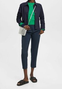 Marinblå kort jacka, grön ribbad tröja, marinblå skräddarsydda byxor och svarta slidesandaler. Silverfärgad crossbody-väska. Släta texturer, figursydd design.