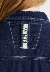 Mørkeblå denimjakke med krave, der har et cremefarvet mærke med "REPLAY" i sort skrift og hvid syning langs kanterne.