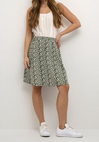 Jupe verte à motifs avec des motifs de feuilles blanches, taille cintrée et silhouette évasée. Portée avec un haut texturé blanc et des baskets blanches.