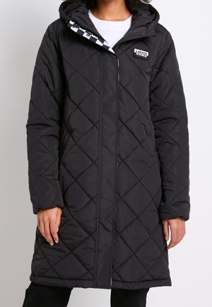 Manteau long noir matelassé à capuche avec un patch logo Vans sur la poitrine, porté par-dessus une chemise blanche.