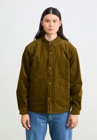 Jachetă de corduroy maro cu guler, închidere cu nasturi în față și două buzunare mari, asortată cu blugi albaștri. Textură netedă, design casual.
