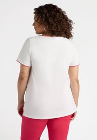 Top blanco de manga corta con ribetes en forma de ola rosa, textura suave y corte relajado, combinado con pantalones rosas. Mostrado desde un ángulo posterior.