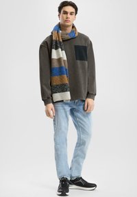 Felpa grigia scura con tasca nera, jeans azzurri chiari, sciarpa a strisce multicolori e scarpe da ginnastica nere indossate da un modello.