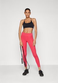 Črn športni nedrček, roza visok pas legice in črne teniske. Ženska drži črna skipping vrv, postavljena na enostavni svetli ozadje.