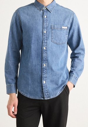 Homme portant une chemise en denim bleu clair boutonnée avec une poche poitrine et un pantalon noir, debout, une main dans la poche.