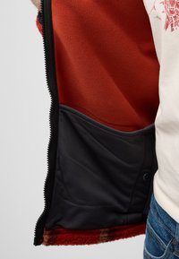 De binnenkant van de fleece-gevoerde jas heeft een rode fleece body, een zwarte nylon voering en een zak met drukknoopsluiting voor opslag. Voorzien van een ritsopening.