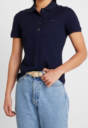 Femme portant un polo bleu marine à trois boutons et un jean bleu clair, ajustant la boucle de ceinture beige d'une main.