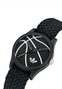 Montre noire avec un bracelet tissé texturé. Le cadran rond présente un motif de basket-ball en noir mat et argent, avec un petit logo en bas.