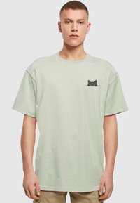 Light green cotton t-shirt with a relaxed fit, featuring a black cat graphic on the left chest. Short sleeves and crew neckline.