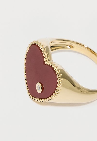 Χρυσό δαχτυλίδι με μεγάλο κόκκινο κεντρικό κόσμημα σε σχήμα καρδιάς, με μικρό πολύτιμο λίθο στο κέντρο και λεπτομέρειες στριφτού σχοινιού κατά μήκος της άκρης.