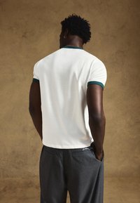 Biała koszulka z krótkim rękawem z ciemnozielonym wykończeniem, zestawiona z szarymi spodniami w cienkie prążki, widziana od tyłu na neutralnym tle.