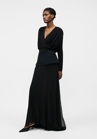 Kobieta o krótkich włosach, ubrana w długą czarną suknię z głębokim dekoltem w serek, dopasowaną talią, długimi rękawami i czarnymi szpiczastymi obcasami, stojąca bokiem.
