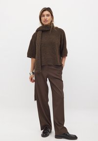 Brauner Strickpullover mit hohem Kragen, halben Ärmeln und einem Fransen-Schal, kombiniert mit weiten braunen Hosen und schwarzen Loafers.