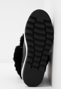 Botte d'hiver noire avec semelle épaisse à motifs et doublure en fourrure douce, positionnée semelle contre un mur d'angle blanc.