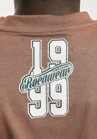 Brown cotton t-shirt featuring a graphic print with "Rocawear" and the numbers "1999" in white and dark green outlined text.