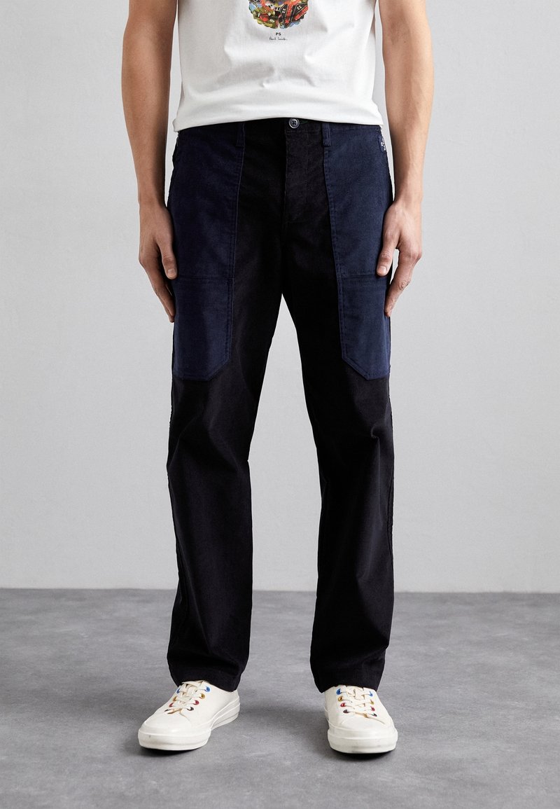 Homme portant un pantalon patchwork noir et bleu foncé, des baskets blanches avec des œillets multicolores et un t-shirt graphique blanc, debout sur un sol gris.