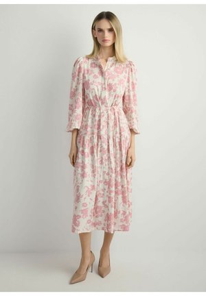 Kobieta ubrana w białą sukienkę midi z różowymi kwiatowymi wzorami, z rękawami za łokieć i beżowe szpilki, stojąca na tle jednolitego jasnego tła.