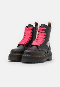 Botas de tobillo de cuero negro con cordones de satén rosa, suela de plataforma, costuras amarillas y un detalle gráfico en el lado exterior. Cierre con cremallera.