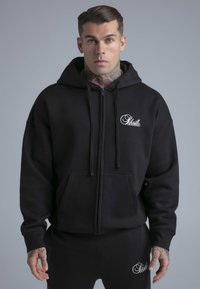 Zwarte zip-up hoodie met voorkantzakken, trekkoorden en geborduurd logo. Zachte stof, losse pasvorm, gecombineerd met bijpassende zwarte joggers.