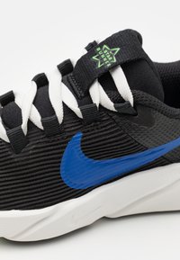 Black sneaker with ridged texture and blue Nike swoosh. Features white laces and a black tag with green "Star Runner" logo. White sole.
