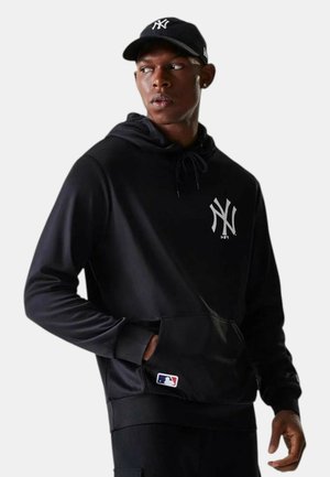 Uomo con cappellino nero dei New York Yankees e felpa con cappuccio con logo MLB, che guarda a sinistra, mani vicino alle tasche della felpa, sfondo grigio chiaro.