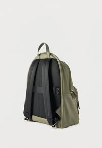 Πράσινο σακίδιο πλάτης με μαύρους επενδεδυμένους ιμάντες ώμου και πίσω πάνελ, ασημένια φερμουάρ και λαβή μεταφοράς στην κορυφή, σε λευκό φόντο.