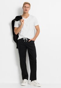Maglietta bianca a V abbinata a pantaloni neri con coulisse elastica. Il modello indossa sneakers bianche e tiene una giacca nera sulle spalle.