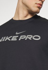 Mustast Nike Pro t-särk lühikeste varrukatega, millel on silmapaistev valge logo ja tekst rinnal. Puuvillane kangas, ümar kaelus.