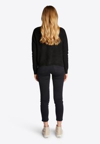 Maglione nero a maglia testurizzata, jeans scuri attillati e sneakers beige alte. I lunghi capelli mossi cadono sulle spalle viste da dietro.