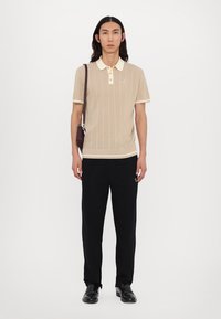 Beige knitted polo shirt with a cream collar and trim, paired with black trousers and black shoes. Minimal branding on the shirt.