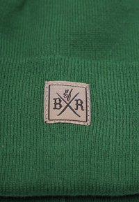 Grön mössa med ribbat mönster, med en rektangulär beige lapp med svart text och korsade linjer. Enkel och funktionell design.
