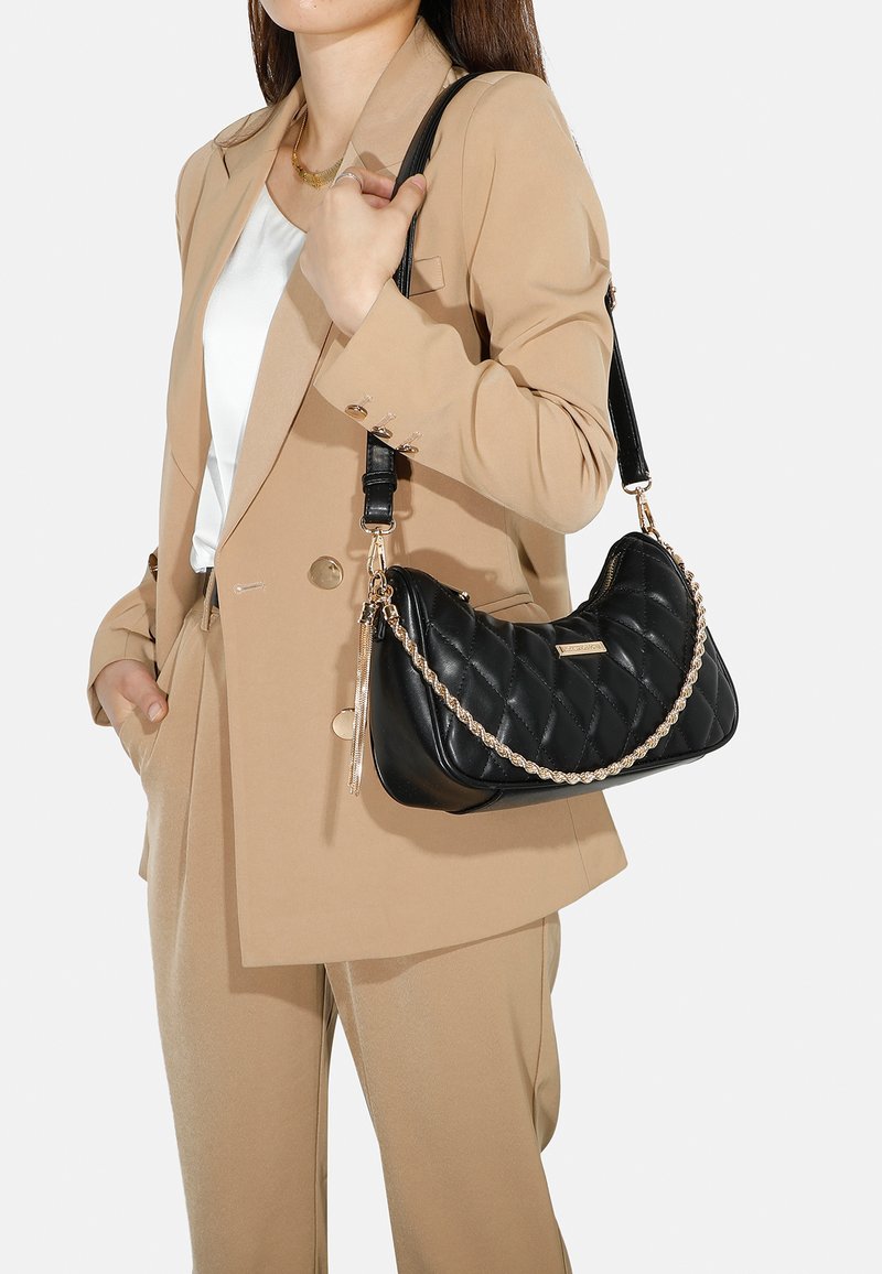 Borsa a tracolla nera trapuntata con una catena d'oro intrecciata, caratterizzata da una texture liscia e dettagli con zip, tenuta da una persona in un completo beige.