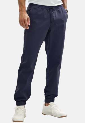 Man wearing navy blue jogger pants with elastic cuffs and waistband, paired with white sneakers and a white shirt.