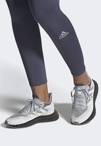 Zapatillas deportivas grises con parte superior de malla texturizada, acentos blancos y una suela distintiva negra impresa en 3D. Combinadas con leggings oscuros.