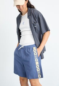Blå shorts med blomsterdetaljer, en casual hvit tanktop og en rutete kortermet skjorte. En beige caps fullfører antrekket.