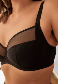 Sort bh med netpaneler, bøjleunderstøttelse og justerbare stropper. Har en glat tekstur og en buet form for pasform og komfort.