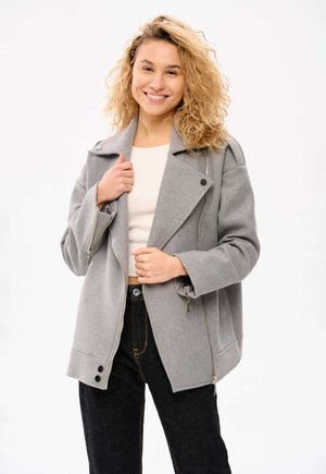 Grau melierte Wolljacke mit Reverskragen, seitlichen Reißverschlüssen und Knopfdetails, kombiniert mit einem hellen Oberteil und dunklen Jeans.