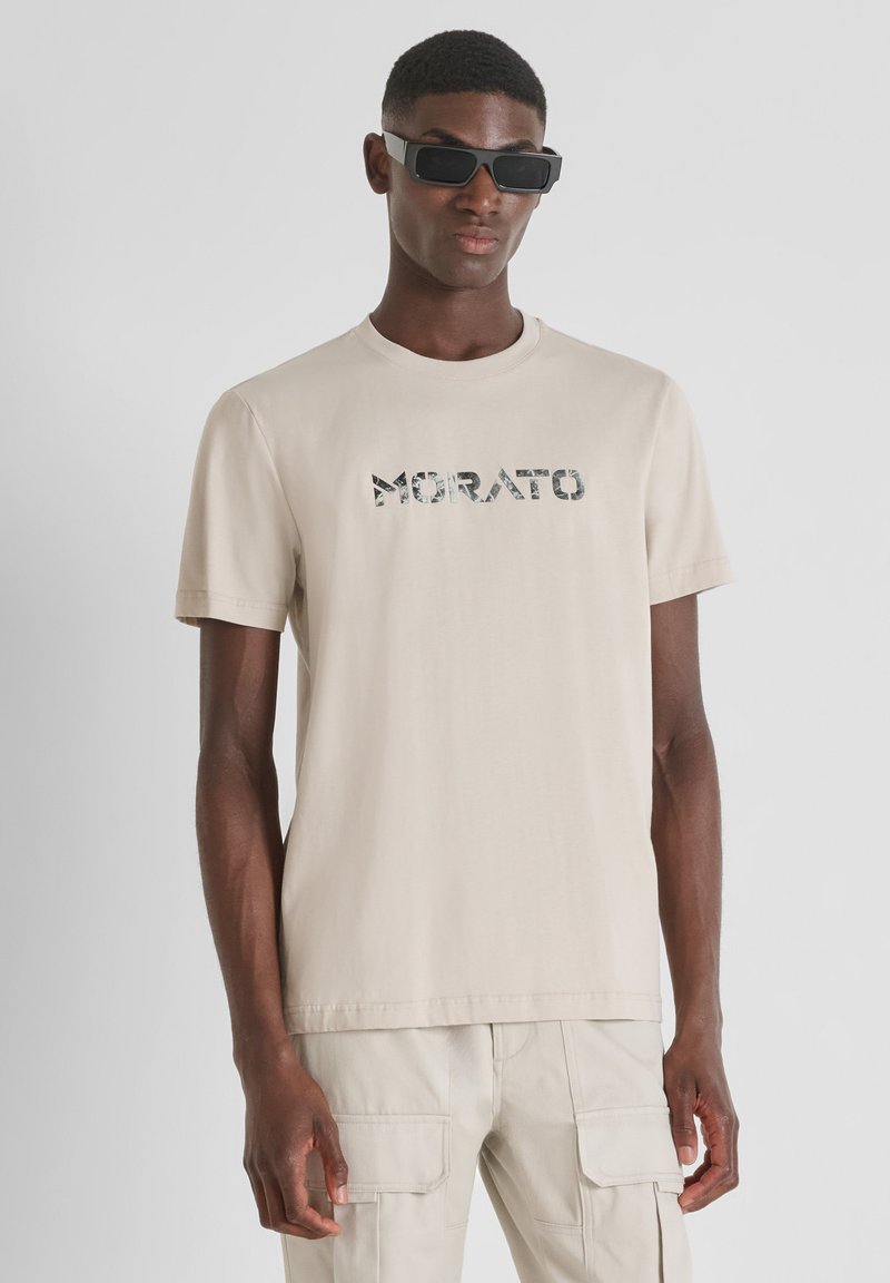 Camiseta de algodón beige con manga corta que presenta la palabra "MORATO" en un patrón de camuflaje. El modelo también lleva gafas de sol rectangulares negras.