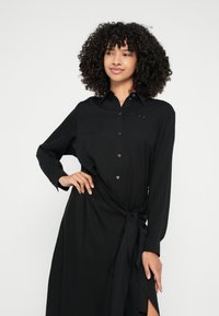 Crna košulja-haljina od glatkog materijala, s dugim rukavima, detaljem vezanja u struku i bočnim prorezom. Suptilni logo na prsima.