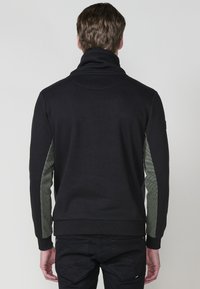 Sudadera negra con cuello alto, mangas con un estampado verde y puños acanalados. Tela suave con una textura lisa, diseñada para la comodidad.