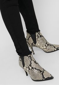 Knöchelhohe Stiefel mit Python-Muster, spitzer Zehenpartie, Stiletto-Absatz und Cutout-Design. Die Stiefel haben eine strukturierte Oberfläche und einen schwarzen Akzent.