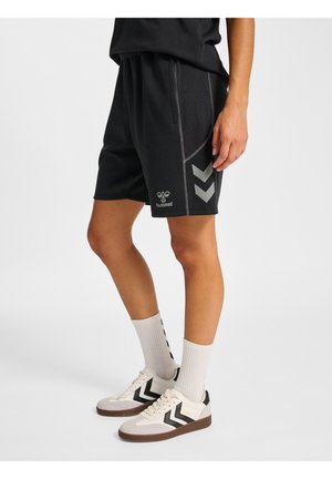 Osoba ubrana w czarne szorty Hummel z odblaskowymi paskami w kształcie chevrona, białe skarpetki i białe buty sportowe z czarnymi detalami w kształcie chevrona, stojąca na szarej podłodze.