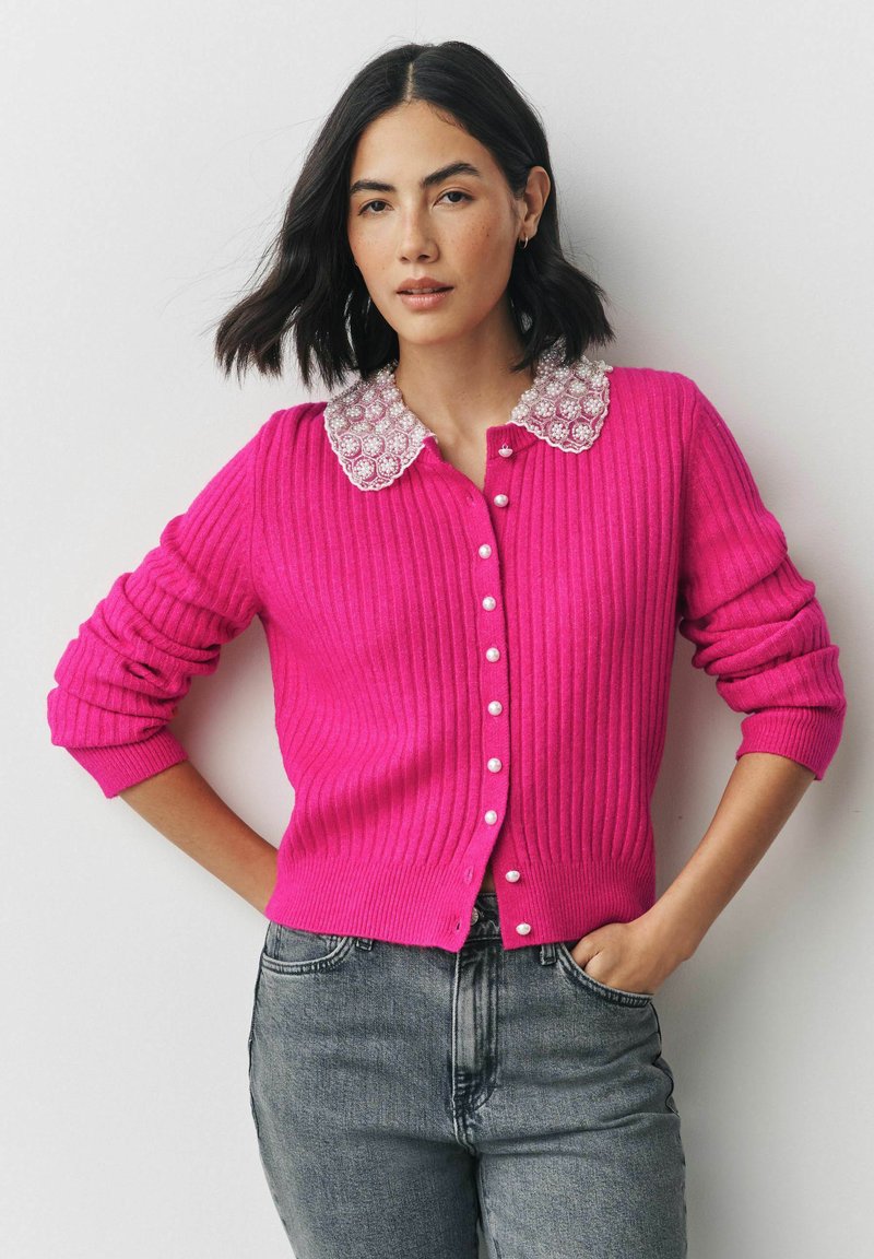 Rosa gerippter Cardigan mit einem weißen verzierten Kragen, ausgestattet mit Perlenknöpfen, langen Ärmeln und einem taillierten Schnitt. Trägt dazu graue Jeans.
